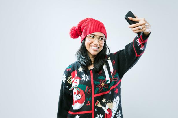 A happy Asian - Pacific Island woman wears an ugly Christmas sweater, having fun during the holiday season.  She takes a self portrait with her smart phone to share on social media.