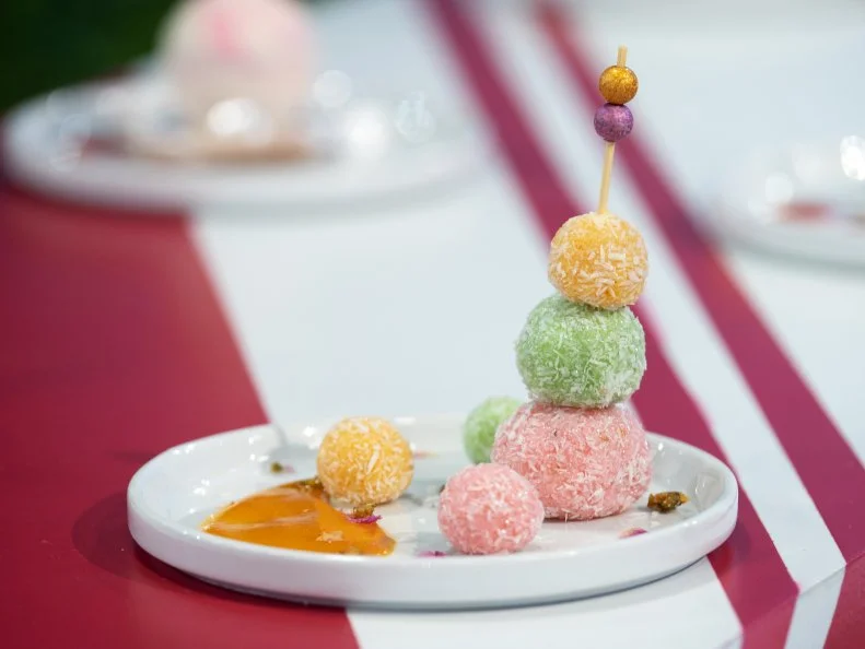 Mini Treat created by Mona Marwaha. It is an Indian-Fusion inspired treat - using a lot of heritage flavors / coconut milk fudge with rose, pistachio, cardamom, mango, saffron, and white chocolate. She made a variety of mini-balls (reminiscent of the land) colored to look like the land and skewered on sticks for judges to eat. There is a mango sauce also on the plate that is part of the flavor combo (so not just decorative), as seen on Candyland, Season 1.