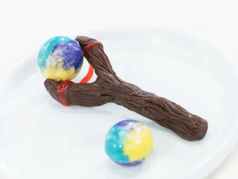 Mini Treat created by Grace Pak is made to look like a "slingshot" but the whole thing is one piece, so the judges can easily pick up the treat as it is practically a treat on a stick. The "Ball" is the edible part, it's a dark chocolate truffle with passion fruit extract and candied almonds. They're encased in a rainbow marbled white chocolate shell, as seen on Candyland, Season 1.