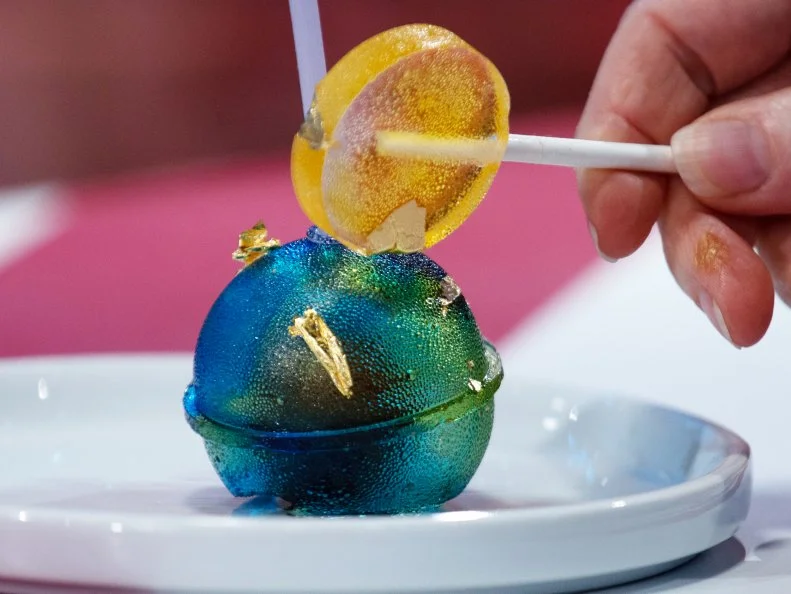 Mini treat created by contestand Jordan Pilarski with the following ingredients: .champagne caramel truffle with a raspberry lime coulis (gooey center) that is covered with a sugar sphere (looks like a big lollipop with edible gold). Served with a smaller yellow lollipop hammer to crack it, as seen on Candyland, Season 1.