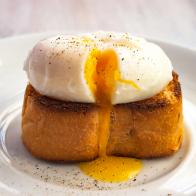Close-up view of tasty poached egg with liquid yolk with fresh green onion on toastbread on white plate on natural white rustic wooden background. Selective soft focus.