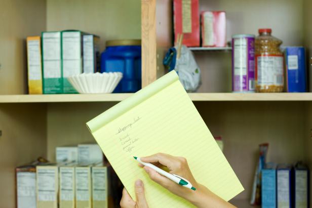 Close-up of a woman's hand writing a shopping list, open pantry in the background. Shallow Depth of Field.