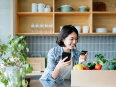 Beautiful smiling young Asian woman grocery shopping online with mobile app device on smartphone and making online payment with her credit card, with a box of colourful and fresh organic groceries on the kitchen counter at home