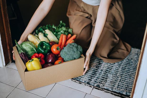 Close up of young Asian woman receiving a box full of colourful and fresh organic groceries ordered online by home doorstep delivery service. Zero waste shopping and home delivery concept