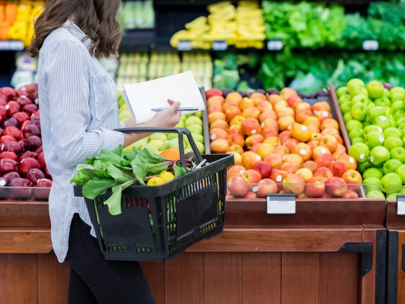 Young woman carries a shopping basket filled with fresh produce. She is shopping for fresh fruit and vegetables in a grocery store.