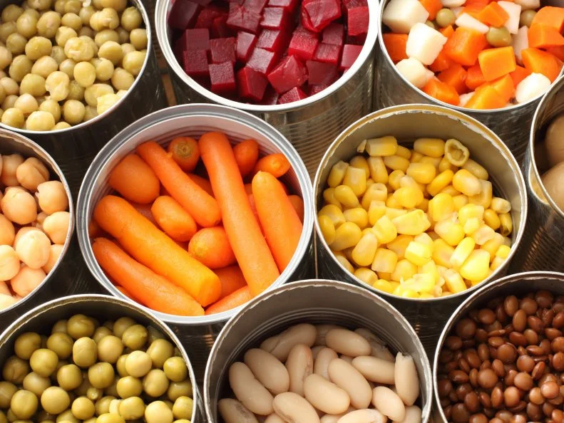 Twelve cans of open vegetables sit next to one another in three rows.  There are peas, carrots, beats, snow peas, corn, chick peas and Lima beans in the cans.