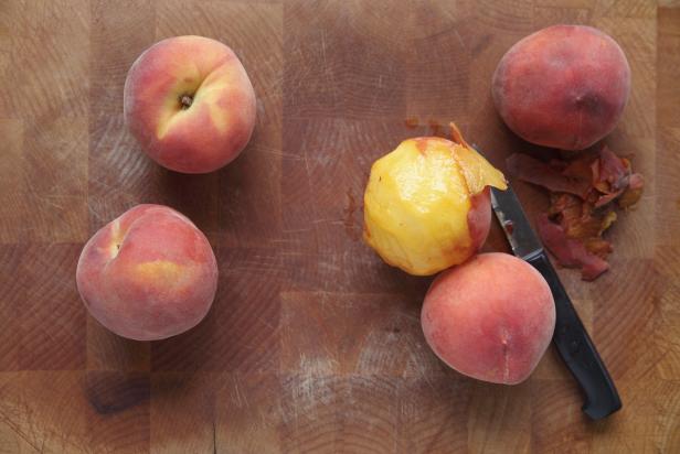 Fresh peaches on a wood cutting board, one being peeled with paring knife