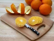 Peeled orange being sliced on chopping board beside coil of orange peel removed from orange. Chopping board on kitchen table with a few more oranges in the background.