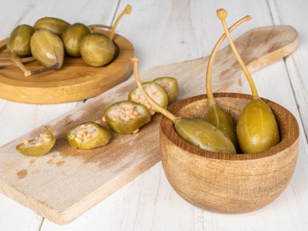 Group of eight whole four slices of pickled green caper on wooden cutting board on round bamboo coaster in bamboo bowl on white wood