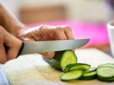 Hands of senior woman cutting cucumber with knife on board in kitchen at home