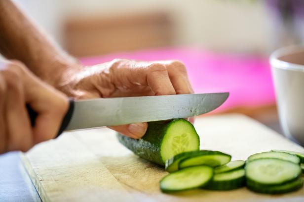 Hands of senior woman cutting cucumber with knife on board in kitchen at home
