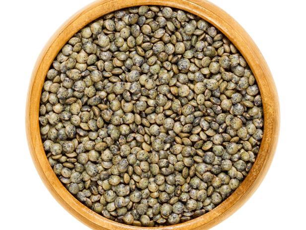 Le Puy green lentils in wooden bowl. Dried small slate-gray seeds of Lens esculenta puyensis from Le Puy in Auvergne, France with unique peppery flavor. Macro food photo close up from above over white