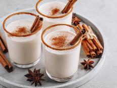 Homemade vanilla Christmas drink Eggnog in glass with grated nutmeg and cinnamon sticks on gray stone background.