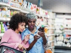 Two African-American women shopping together in a supermarket. The younger one, in her 20s, is carrying a shopping basket. The older one, a senior woman in her 60s, is holding a bottle and they are reading the nutrition label.