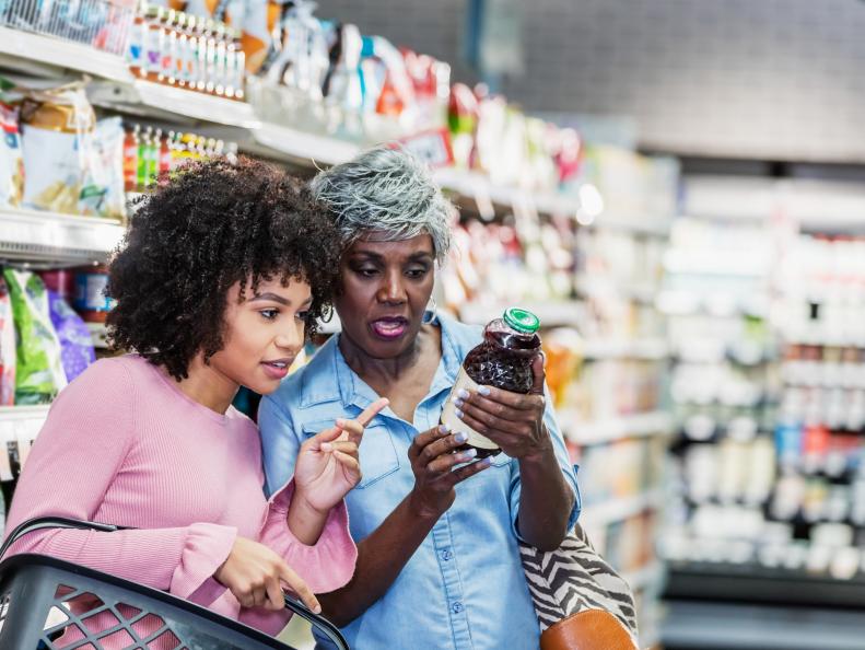 Two African-American women shopping together in a supermarket. The younger one, in her 20s, is carrying a shopping basket. The older one, a senior woman in her 60s, is holding a bottle and they are reading the nutrition label.
