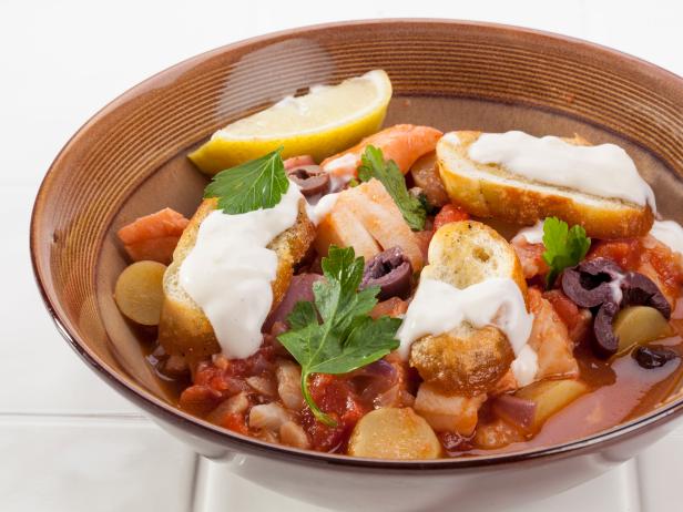 Fish stew in a brown bowl with parsley, olives, potato, salmon, cod and tomatoes. Topped with toasted bread and aioli.