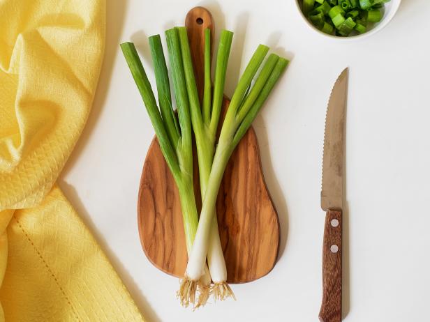 Fresh green onions on a wooden cutting board with a napkin and a knife on a white table.
