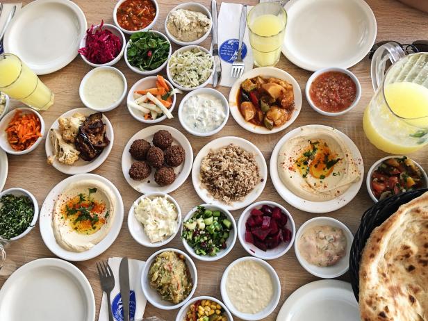 A table with Israel Appetizers - Hummus, Falafel, Tahini, Salads, Guacamole, Couscous, Pita Bread, Tabbouleh, Tzatziki, Rice, Feta, Meze, Cabbage, Eggplant, Corn, Carrot, Zucchini. Fresh juice.