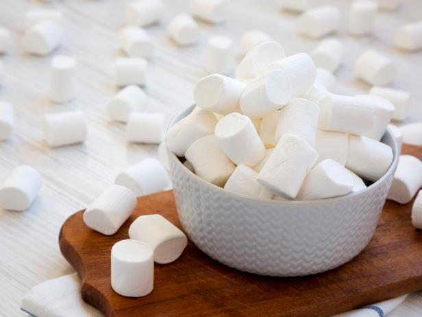 White fluffy marshmallows in a bowl on rustic wooden board over white wooden surface, side view. Close-up.