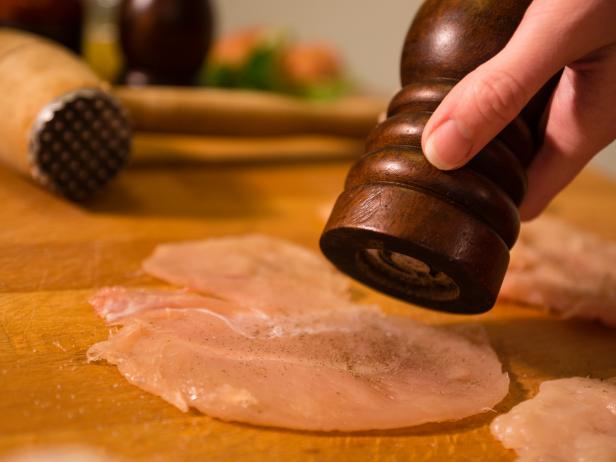 Close-up of chicken breast being seasoned with pepper using a wood pepper grinder.