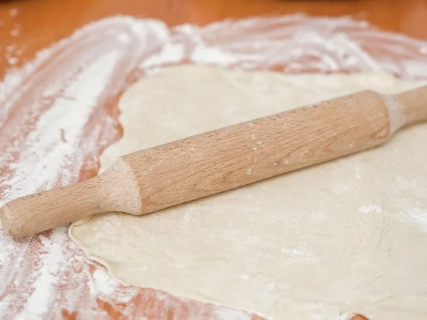 A wooden rolling pin rolls the dough sprinkled with flour. Homemade cake. Butter dough. Space for text. Mistress in the kitchen.