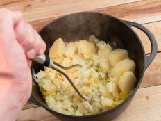 Pressure-cooked potatoes and leeks transferred into black cast iron cooking pan and being mashed. Slightly out of focus human hand holding potato masher. Wooden plank. High point of view.