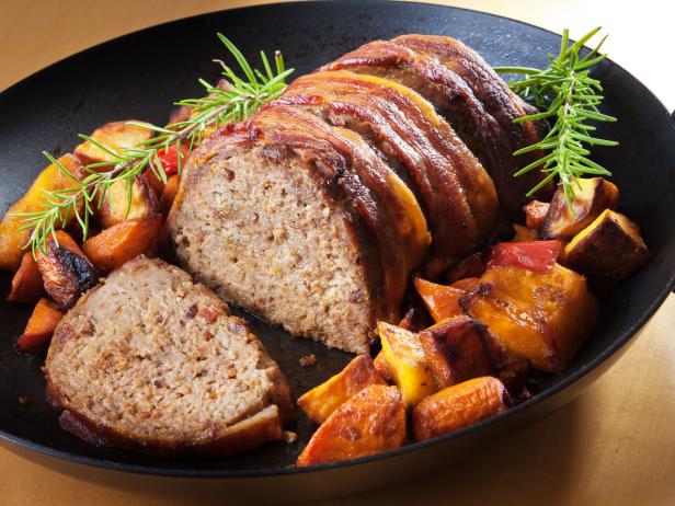 Meatloaf, a favorite and popular American main dish, roasted with bacon covering and accompanied by roasted vegetable such as carrots, squash, and potatoes, garnished with sprigs of rosemary. Presented on a black iron roasting pan.