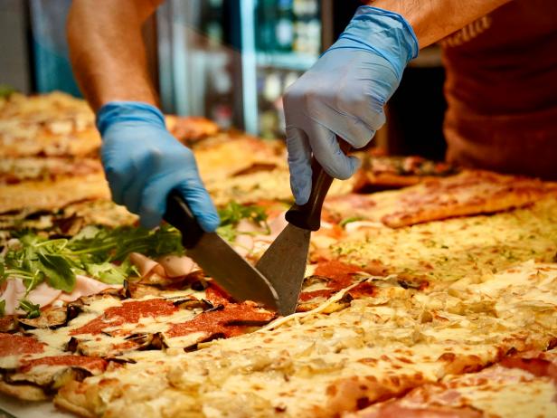 An employee at La Renella cuts a piece of pizza for a customer, as seen on Bobby & Giada In Italy, Season 1.