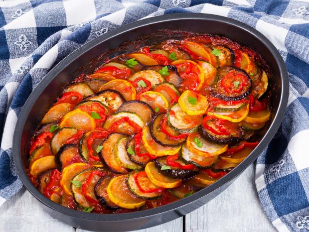 Layered ratatouille in a baking dish, slices of zucchini, red bell pepper, chili, yellow squash, eggplant, olive oil, parsley and garlic on a white background, close-up