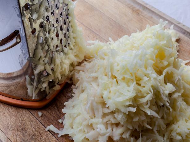 Grated potato with metal standing grater on wooden cutting board from high angle closeup