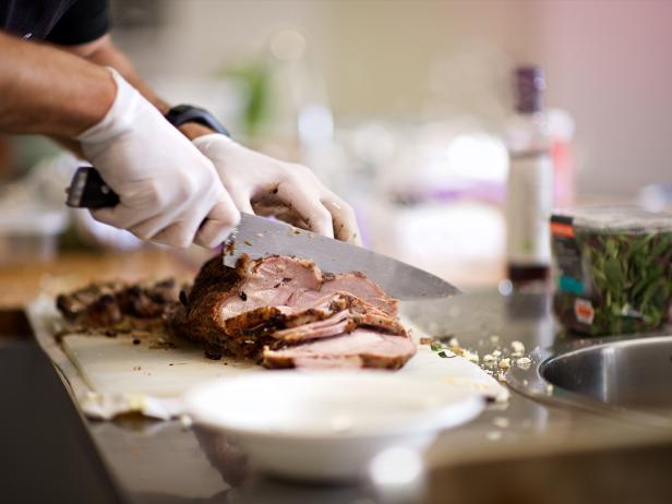 A Caucasian hand with white kitchen surgical gloves Cutting a beef roast with a large knife in a kitchen