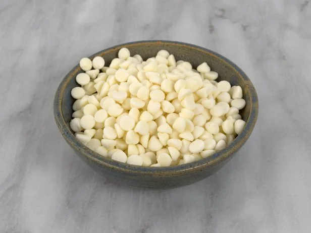 White chocolate baking chips in an old stoneware bowl atop a marble cutting board.