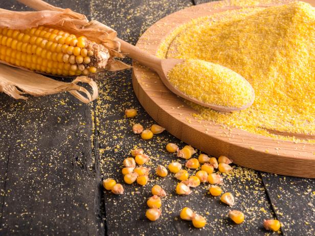 Food ingredients theme image with a corn cob, grains and corn flour on a wooden trencher and spoon, on a rustic wooden table.