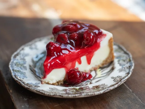 A New York Cheesecake with cherries sits on a wood table. The cake is served on a single antique plate.