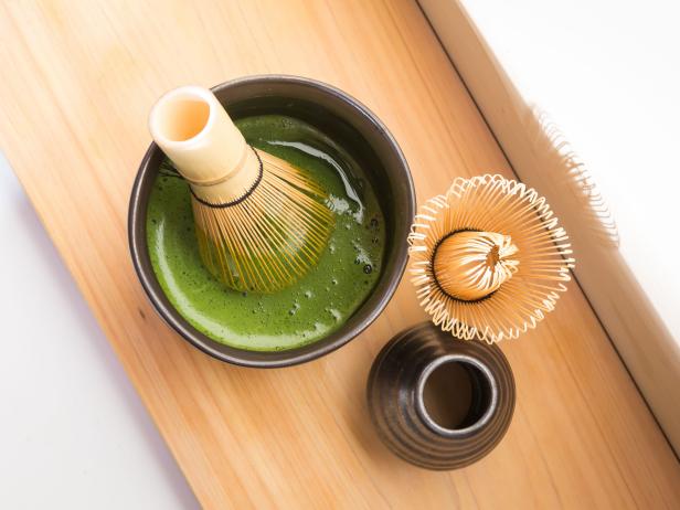 Mixed organic green Matcha in a back bowl with a traditional Matcha Whisk.  An unused whisk can be seen at the edge of the frame.  There is a bamboo wood panel below the bowl. Angle of the photo is overhead.