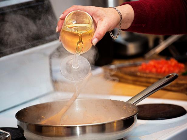Cook pours wite wine into food in a fryingpan on the stove