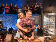 Co-host Damaris Phillips wraps up chef Bobby Flay as she plays the ukulele, as seen on Beat Bobby Flay, Season 26.