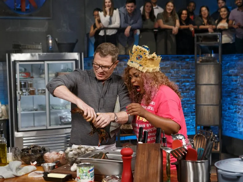 Co-host Sunny Anderson wears contender Francesco Buitoni's pasta crown and scepter as she checks in on chef Bobby Flay while he cooks Seafood Fra Diavolo during round two, as seen on Beat Bobby Flay, Season 21.
