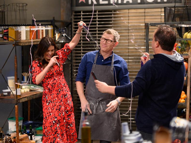 Co-hosts Katie Lee and Josh Capon spray host Bobby Flay with silly string as he races to finish his pasta ragu dish, as seen on Beat Bobby Flay, Season 25.