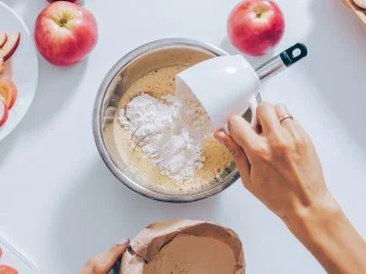 Top view woman adding flour into dough to prepare apple pie. Flat lay composition of kitchen table with ingredients for cooking seasonal fruit cake.