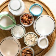 Variety of non-dairy vegan lactose free nuts and grain milk almond, hazelnut, coconut, rice, oat in glass bottles and ceramic jugs with ingredients above over white marble background. Flat lay, space
