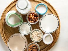 Variety of non-dairy vegan lactose free nuts and grain milk almond, hazelnut, coconut, rice, oat in glass bottles and ceramic jugs with ingredients above over white marble background. Flat lay, space