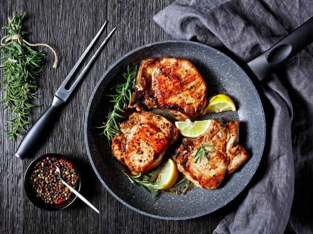 roasted pork chops with rosemary sprigs and lemon slices in a skillet on a wooden table, horizontal view from above, flat lay