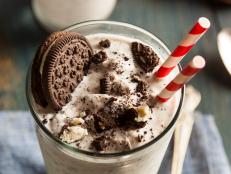 A cookies and cream milkshake sits on a wooden table in a tall glass with a spoon beside it.  There is a large chocolate sandwich cookie in the milkshake along with crumbled chocolate cookies.  There are two old-fashioned red-and-white straws standing in the glass.