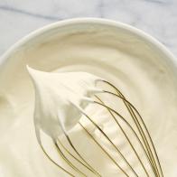 Close up view of whipped cream on whisk with cream in background.