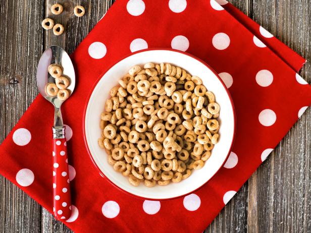 A bowl of cereal and a red polka dot napkin on a wooden table.