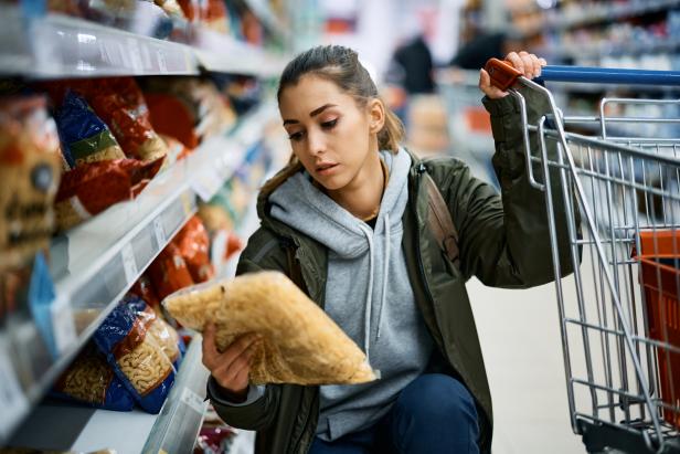 Young woman looking at the price on pasta package while shopping in grocery store.