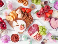 Mother's Day theme charcuterie table scene against a white wood background. Variety of cheeses, meats, fruit and sweets. Top view.