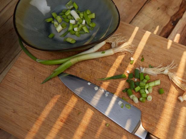 Chopping scallions. Chopped scallions in colourful crockery.  Kitchen knife. Bamboo cutting board on wooden plank. Light effect. High point of view.