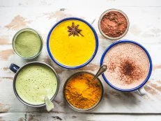 A Matcha Latte, a Golden Milk Latte and a Raw Cacao Hot Chocolate assembled in a cluster on a rustic white wooden table top, with jars of the ingredients gathered around.